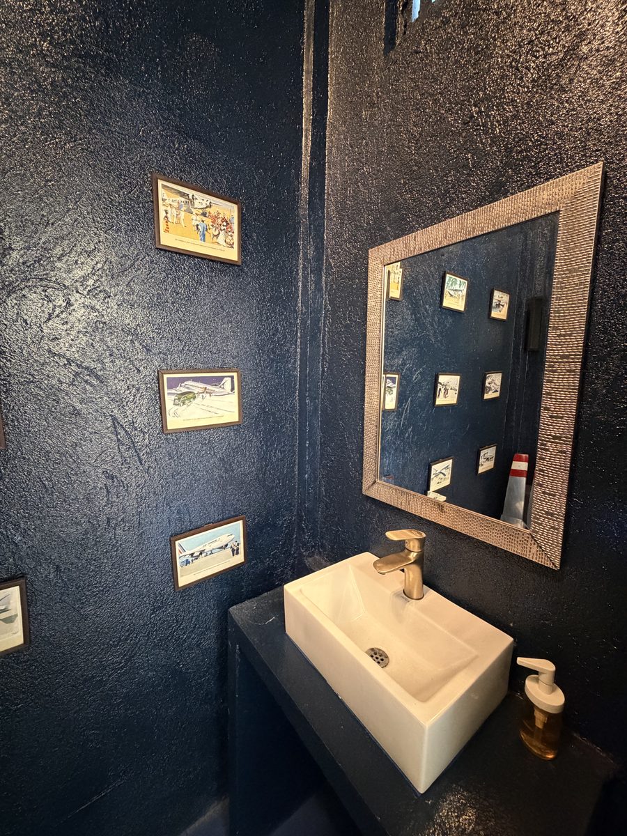 Baño con pared azul profundo, lavamanos blanco y espejo de marco tejido
